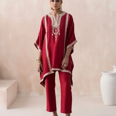 A clean, full-length lookbook image styled for Saisha's 'Firdaus' collection. The focus is on the sharp silhouette of this red kaftan set, with minimal props and a neutral background to make the intricate embroidery and vibrant color stand out.