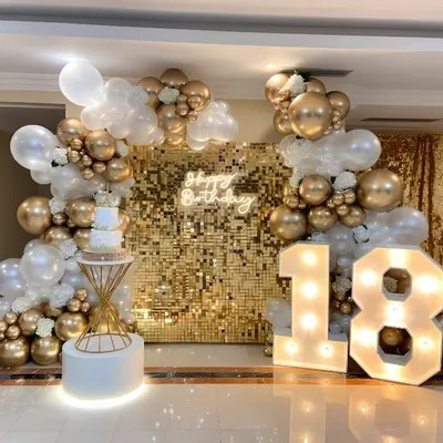 An elegant 18th birthday party setup featuring a gold sequin wall and a 'Happy Birthday' neon sign. The decor is completed with a white and gold organic balloon garland and large light-up marquee numbers.