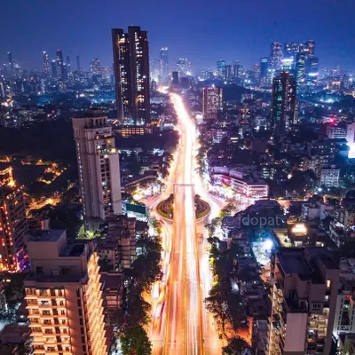 A high-angle, long-exposure drone shot of a major traffic circle in Mumbai at night, with light trails creating a sense of organized chaos.