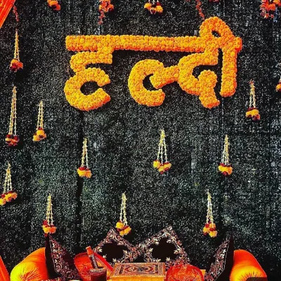 This is a close-up of our signature Haldi ceremony backdrop, a highly requested design for its traditional beauty. We crafted the word "Haldi" in Hindi using dense, fresh marigold flowers, set against a dark green foliage wall. The design is completed with delicate hanging floral tassels, creating a rich, textured look that is perfect for photos.