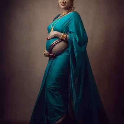 A full-length portrait showcasing the grace of a mother-to-be in a beautiful teal saree. The studio lighting is carefully set to highlight the texture of the fabric and the gentle curve of the baby bump.