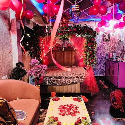 This is a wide view of a complete romantic room decoration. The setup includes a bed canopy of red roses, fairy lights, red balloons on the ceiling, and a table set with candles and rose petals for a memorable surprise.