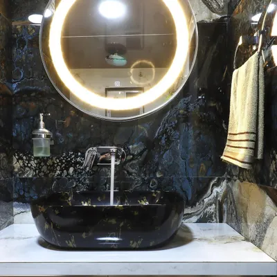 A luxurious powder room featuring a statement black and gold vessel sink. The dramatic black marble wall and backlit circular mirror create a bold and sophisticated atmosphere.