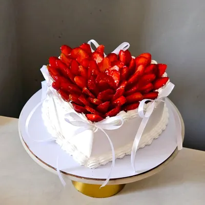 A heart-shaped chocolate ganache cake, generously topped with a beautiful arrangement of fresh, sliced strawberries. The white ribbons add a delicate touch, making it a perfect cake for an anniversary or special treat.