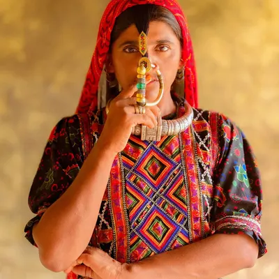 A powerful portrait of a Dhaneta Jat woman from Kutch, her face partially obscured by the large, traditional nose ring she holds. Her direct gaze is captivating and full of strength.