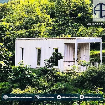 This minimalist container home is nestled in a lush, green landscape, offering an off-the-grid living experience. Its simple white exterior contrasts beautifully with the natural surroundings.