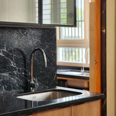 A close-up of a kitchen sink area, showcasing the rich texture of the black marble backsplash against the warm wooden cabinetry. The fluted glass of the overhead cabinet adds a layer of translucency and refinement.