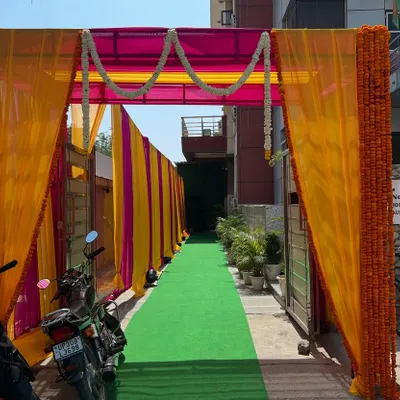A vibrant entrance for a daytime event, created with yellow and pink drapes and marigold garlands over a green carpet walkway.