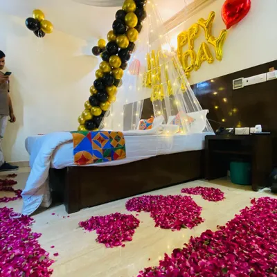 A luxurious birthday room decoration featuring a path of rose petals leading to the bed. The decor includes a canopy with fairy lights, a black and gold balloon garland, and "Happy Birthday" foil balloons.