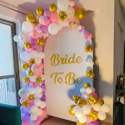 A lovely "Bride to Be" backdrop for a home party. This setup features a white arched panel with gold lettering, framed by a colorful balloon garland in shades of pink, purple, white, and gold.