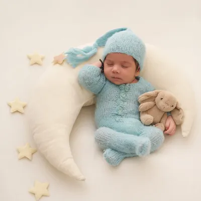 A baby in a cozy blue sleeper and hat cuddles a bunny while sleeping on a soft crescent moon pillow, surrounded by stars.