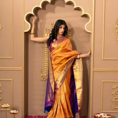 A full-length shot of a model in a vibrant orange and purple silk saree from Kumaran Silks. The grand, traditional backdrop complements the festive and formal nature of the attire.