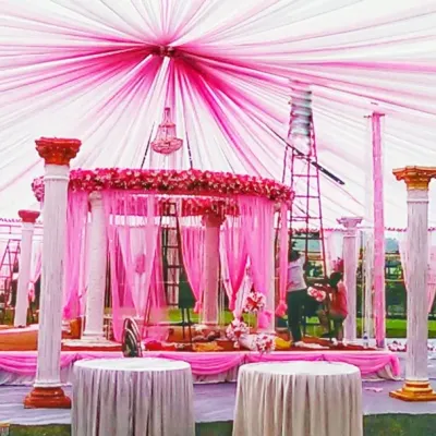 This image shows a wedding pandal during setup, highlighting the structural work involved. The design features decorative pillars and an elaborate pink and white fabric ceiling, creating a grand mandap for the ceremony.