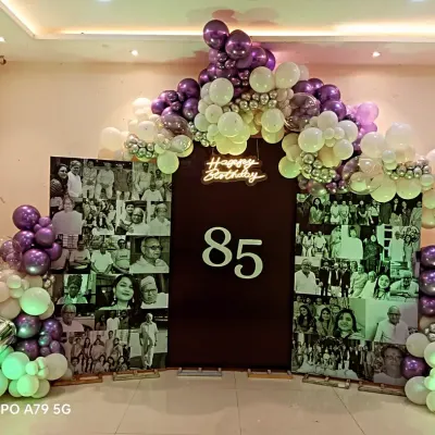 A heartfelt tribute for an 85th birthday celebration. This large backdrop features a collage of black and white photos, framed by a beautiful balloon arch in purple, silver, and white.