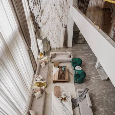 An aerial view of this double-height living area, showcasing the grandeur and light we aimed to achieve. The layout is planned for easy conversation, with distinct seating groups anchored by marble coffee tables and emerald green accent chairs, all under a cascading statement chandelier.