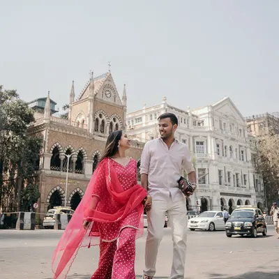 A classic Mumbai pre-wedding shot, with the couple taking a stroll against the backdrop of historic architecture. Her vibrant pink suit adds a beautiful pop of color to the city scene.