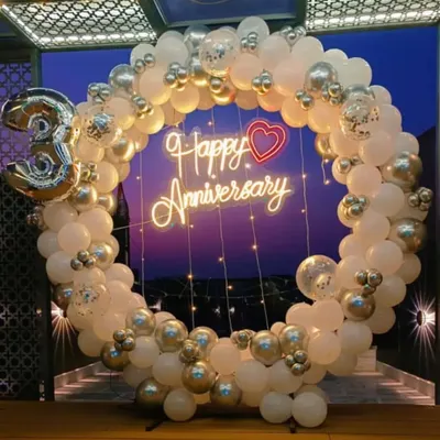 A beautiful anniversary backdrop featuring a full circle balloon arch in white and silver chrome. The design is completed with a "Happy Anniversary" neon sign and a large number "3" balloon for a third anniversary.