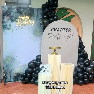 A sophisticated "Chapter Twenty-eight" birthday setup. This features a marble-effect backdrop, a modern arch panel, black balloons, and elegant white plinths.