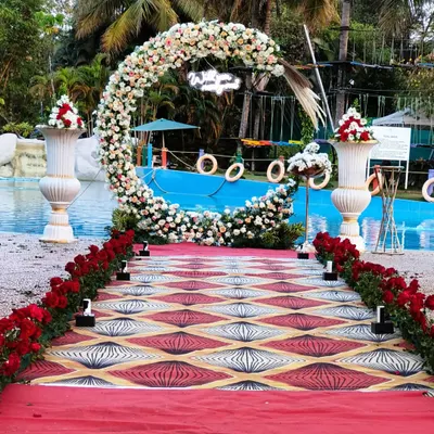 This poolside entrance features a circular floral arch with a "Welcome" sign, leading guests down a custom-printed aisle runner. The red rose border adds a classic touch to this modern outdoor wedding setup.