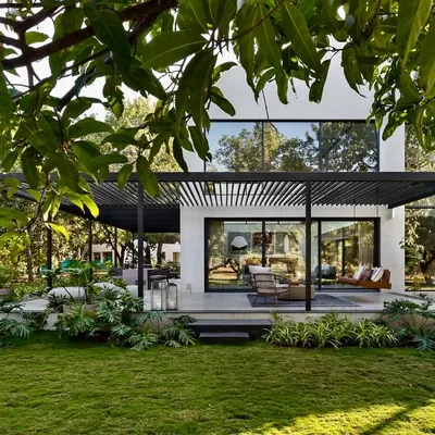 The exterior of the Mango Orchard House shows how the structure opens up to the landscape. A large, shaded terrace extends the living space into the garden, creating a perfect setting for outdoor living while being sheltered by a modern pergola.
