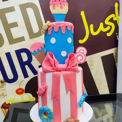 A playful two-tier custom cake designed for a fun-filled party. The bottom tier features pink and white stripes with a large fondant bow, while the top blue polka-dotted tier is topped with a melting ice cream cone, lollipops, and other candy decorations.