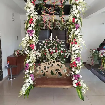 A beautifully decorated traditional wooden swing (jhoola) for a housewarming or pre-wedding ceremony at home. The swing is heavily adorned with vertical garlands of white, red, and purple flowers and exotic leaves.