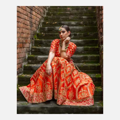 A full-length portrait of a model in a vibrant orange and gold embroidered lehenga, seated on rustic stone steps. This shot highlights my ability to capture the grandeur of Indian bridal couture in an outdoor, editorial setting.