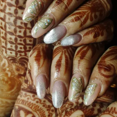 A close-up of long, almond-shaped bridal nail extensions designed to complement detailed Mehendi. The nails feature a shimmery silver polish and hand-painted leafy green motifs with glitter accents, creating a cohesive look with the henna art.