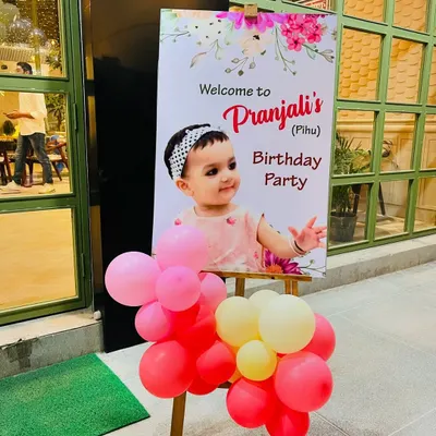 A personalized welcome sign on an easel for a first birthday party, decorated with a small balloon cluster to greet guests.