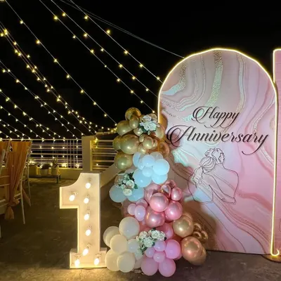 A beautiful first anniversary celebration on a terrace under string lights. The setup features a custom arched backdrop with a marble texture, a balloon arrangement, and a large light-up number '1'.