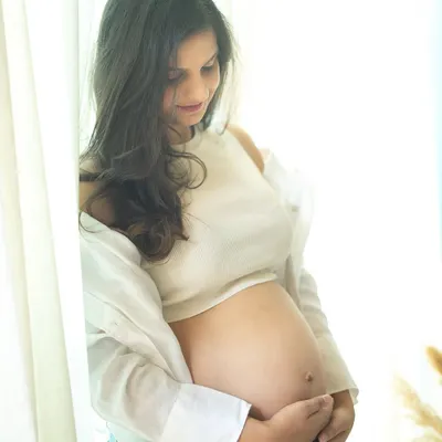 A serene and beautiful solo portrait. The soft window light illuminates the mom-to-be, who is dressed in a casual crop top and jeans, creating a gentle and authentic image.