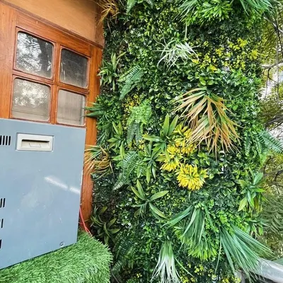 A dense and textured artificial green wall installed next to a wooden window frame. The variety of faux ferns, leaves, and yellow accents creates a realistic, jungle-like effect.