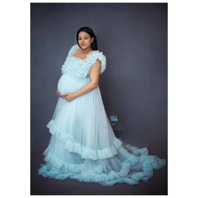 A stunning solo maternity portrait showcasing the mother-to-be in a flowing, light blue ruffled gown against a classic studio backdrop.