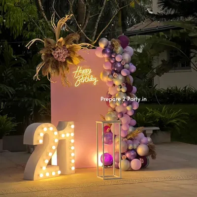 A chic and modern 21st birthday setup in an outdoor garden setting. This design features large light-up marquee numbers, a stylish pink backdrop, and a beautiful balloon garland with pampas grass and floral details.
