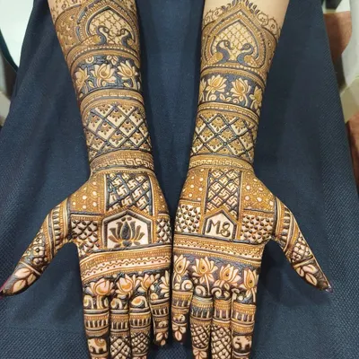 A classic full-hand bridal mehndi design, rich with traditional patterns. It features beautiful lotus flowers, peacocks, and intricate checkwork, with a personalized initial monogram on the palm.