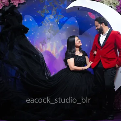 A dramatic pre-wedding studio shot featuring a flowing black trail gown and a bold red blazer. The moon prop and floral background create a fantasy-like, romantic atmosphere.