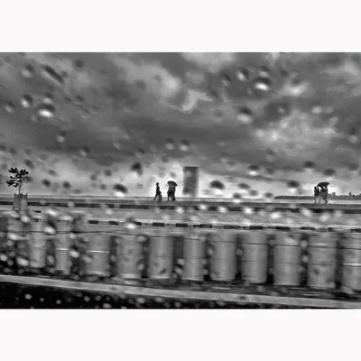 A moody, atmospheric scene of a rainy day, viewed through a water-streaked window. The motion blur and droplets create a painterly effect, turning a mundane moment into a piece of evocative black and white street photography.