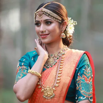A beautiful close-up of a South Indian bridal look. The focus is on the detailed makeup, the traditional temple jewellery, and the rich colors of the orange and teal silk saree.
