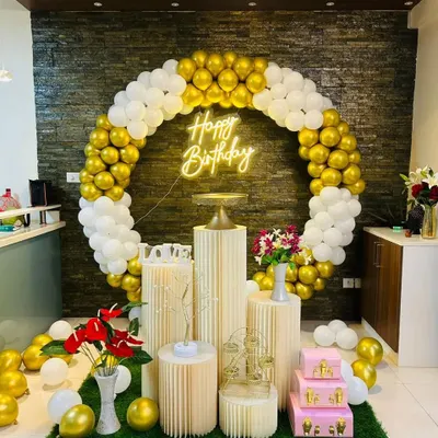 A classic and elegant birthday setup with a full circular balloon arch in gold and white. The decoration is placed on an artificial grass mat and includes pedestals, floral arrangements, and a 'Happy Birthday' neon sign.