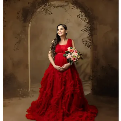 A full-length portrait of a mother-to-be in a stunning red ruffled gown, holding a bouquet of flowers against a rustic archway backdrop. This is a perfect example of our fine art maternity portraiture.