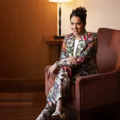 A full-length portrait of Saina Nehwal in a stylish floral pantsuit. The relaxed, seated pose in a well-lit room creates a look that is both professional and effortlessly chic.