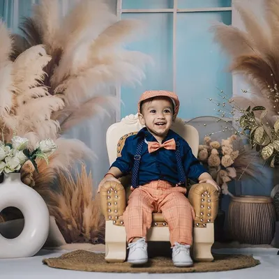 A dapper young boy in a bow tie and cap smiles from his miniature armchair. This studio portrait uses a beautiful boho-themed backdrop with pampas grass for a stylish and warm feel.