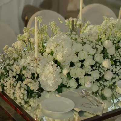 A mirror-top table reflects a lush arrangement of white hydrangeas, roses, and baby's breath, paired with elegant taper candles for a timeless and romantic look.