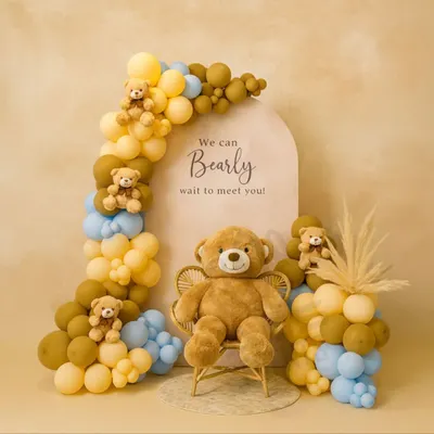 The classic 'We can Bearly wait' baby shower theme. This setup features a charming balloon arch in pastel yellow, blue, and brown, with small teddy bears nestled in the balloons and a large one seated on a wicker chair.