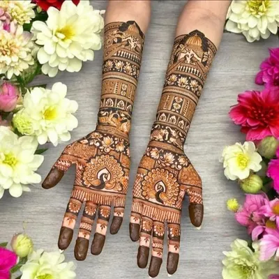 This beautifully staged photo shows a classic Rajasthani design with symmetrical peacocks on the palms and intricate architectural details on the arms, surrounded by fresh flowers.