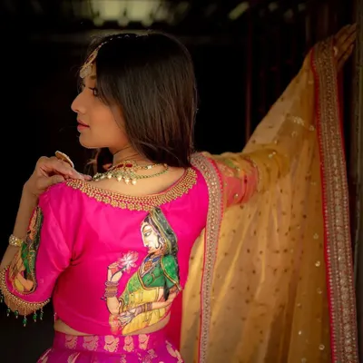 This bright pink raw silk blouse features a beautiful, hand-painted depiction of a traditional figure, making it a wearable piece of art. The delicate gold embroidery along the neckline adds a touch of festive elegance.