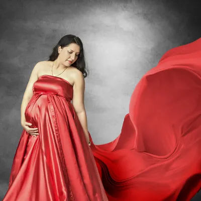 The power of a classic red gown. This studio portrait uses dramatic fabric and soft lighting to create a timeless and elegant image, focusing on the mother's serene expression as she holds her baby bump.
