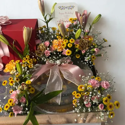 A beautiful birthday hamper where gifts and fresh flowers come together to create a stunning display. This arrangement features a vibrant mix of lilies and carnations alongside carefully selected presents, all tied together with delicate pink ribbons.