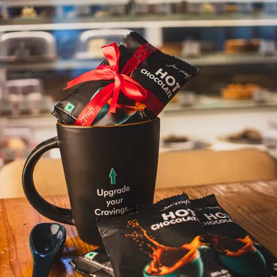 Our Hot Chocolate Mix packs sit inside and beside a black mug, with a red ribbon around the sachets.