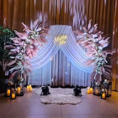 The perfect setting to ask the big question. This 'Marry Me' proposal setup features a neon sign, a beautiful arch with pampas grass, and a floor covered in candles and a soft rug.